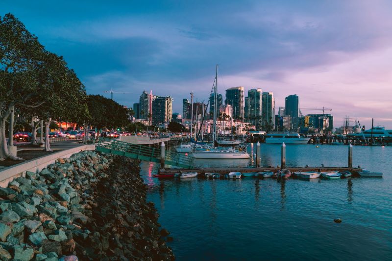 Boats float while docked at the marina at sunset, with the San Diego skyline glittering in the background.