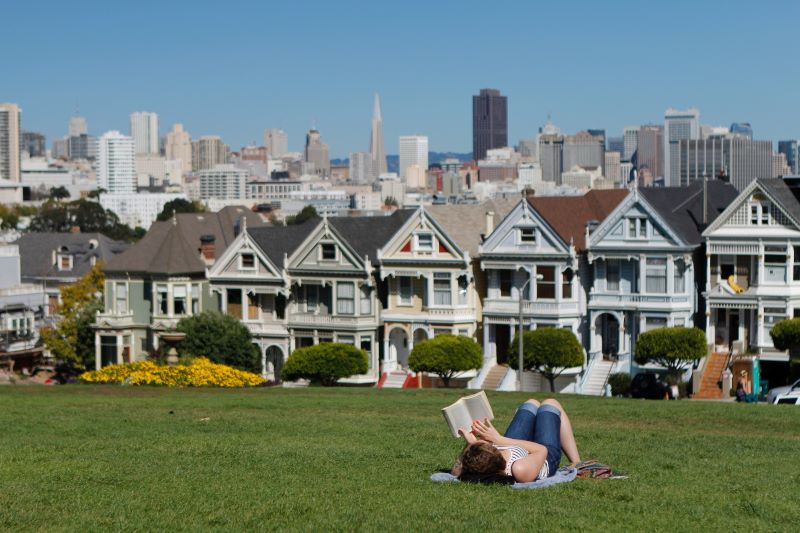A woman reads a book on a grassy hill overlooking the historic painted ladies and greater San Francisco skyline.