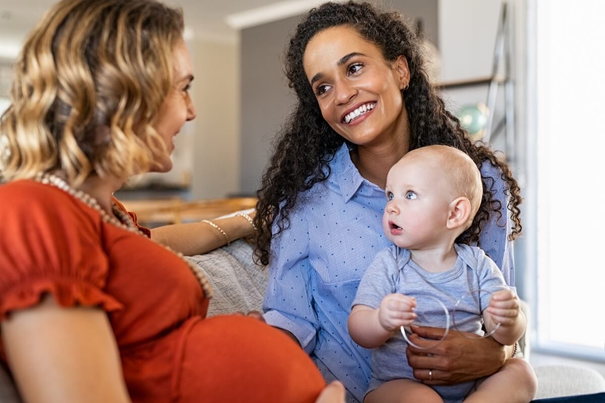 Two multiethnic women smiling at each other, one is pregnant and the other is holding an infant on her lap. Image for the Best Cities for New Moms.