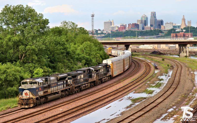 A train passes under a bridge with the Kansas City skyline in the distance