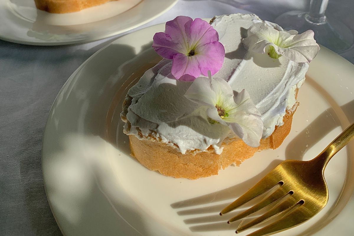 A piece of bread with cream cheese and flowers on top, on a plate with a gold fork