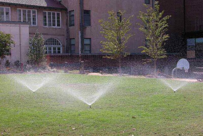 three sprinklers watering a lawn with building and trees in the background