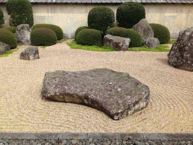 A large boulder in a Japanese Zen garden with bushes in the background