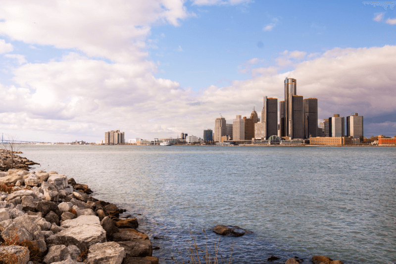 A lake shimmers in front of the Detroit skyline as clouds creep overhead.