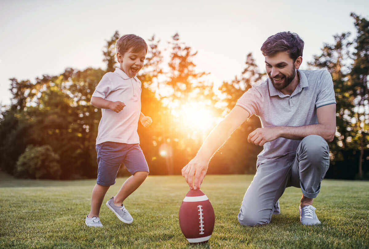 Handsome dad with his little cute sun are having fun and playing American football on green grassy lawn