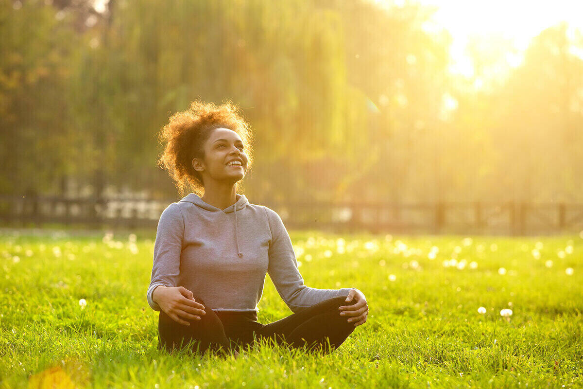 A woman smiles while having a meditative moment sitting cross-legged in the grass.