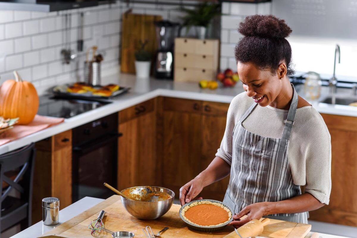 Woman making pumpkin pie on her kitchen island in the foreground; a pumpkin sits on the kitchen counter in the background