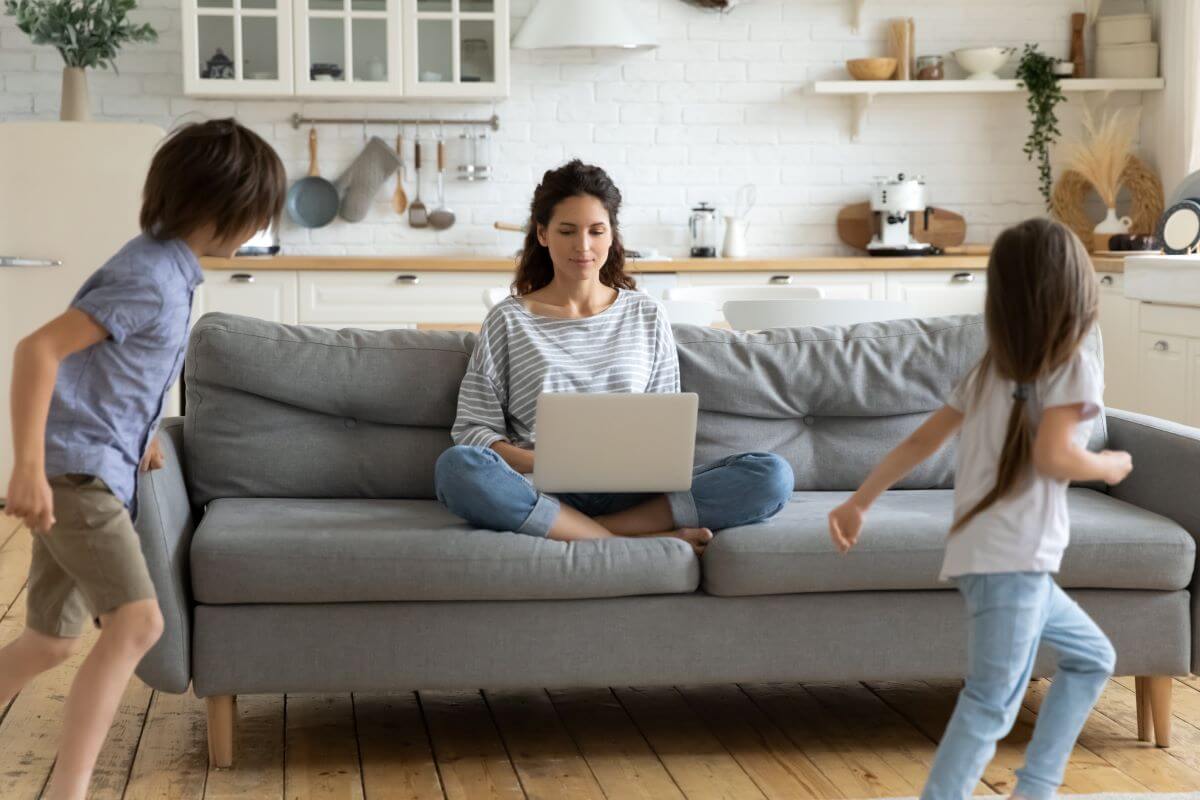 A mom sits on the couch, trying to work on her laptop while her children run around