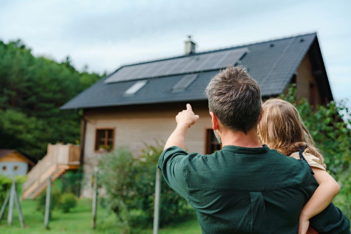 Dad carrying his daughter in the foreground, pointing to solar panels on the roof of their house