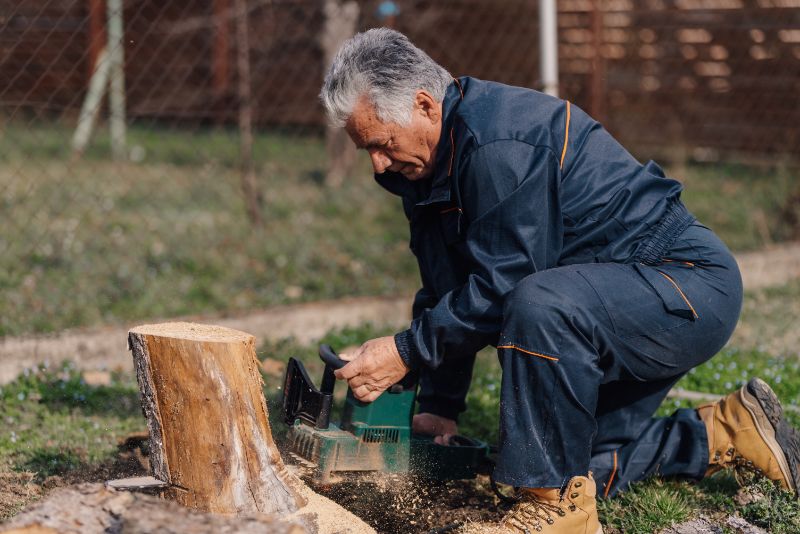 An elderly man in work overalls uses a power tool to grind and remove a tree stump outdoors in a yard. 