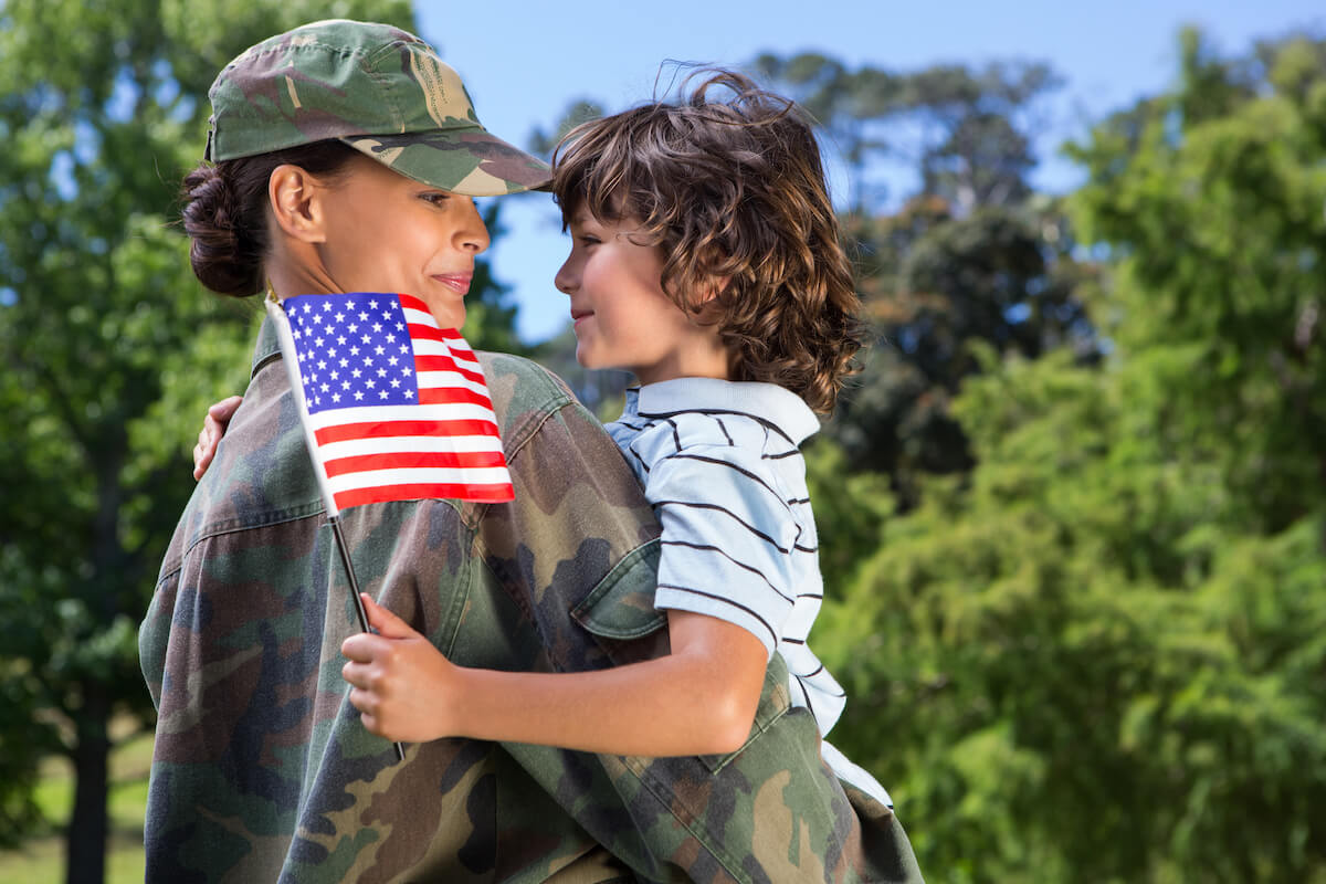 Mother soldier smiling and holding son who is wearing a striped shirt and waving an American flag outside