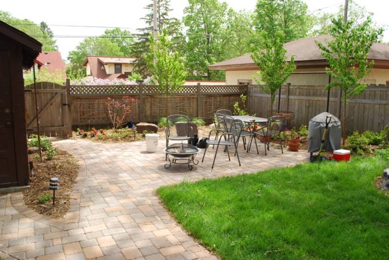 Backyard patio with interlocking pavers, outdoor furniture, and a barbecue grill, surrounded by small garden beds and a patch of lawn.