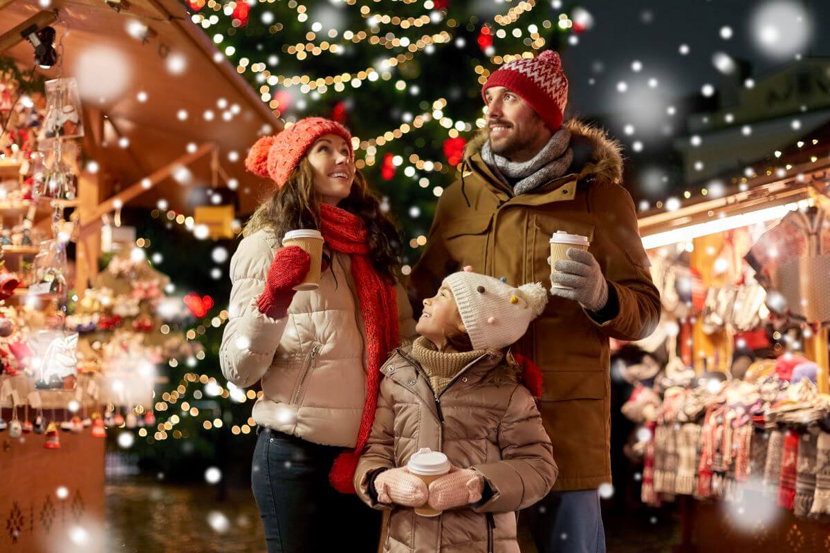 Family looking up at snow and clutching warm beverages at a Christmas market