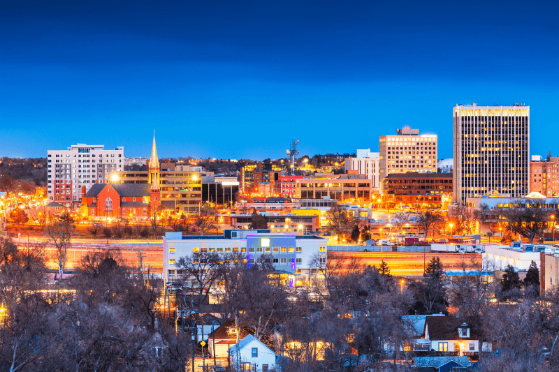 Trees and homes stand amid a background of commercial buildings in Colorado Springs, Colorado, at twilight.