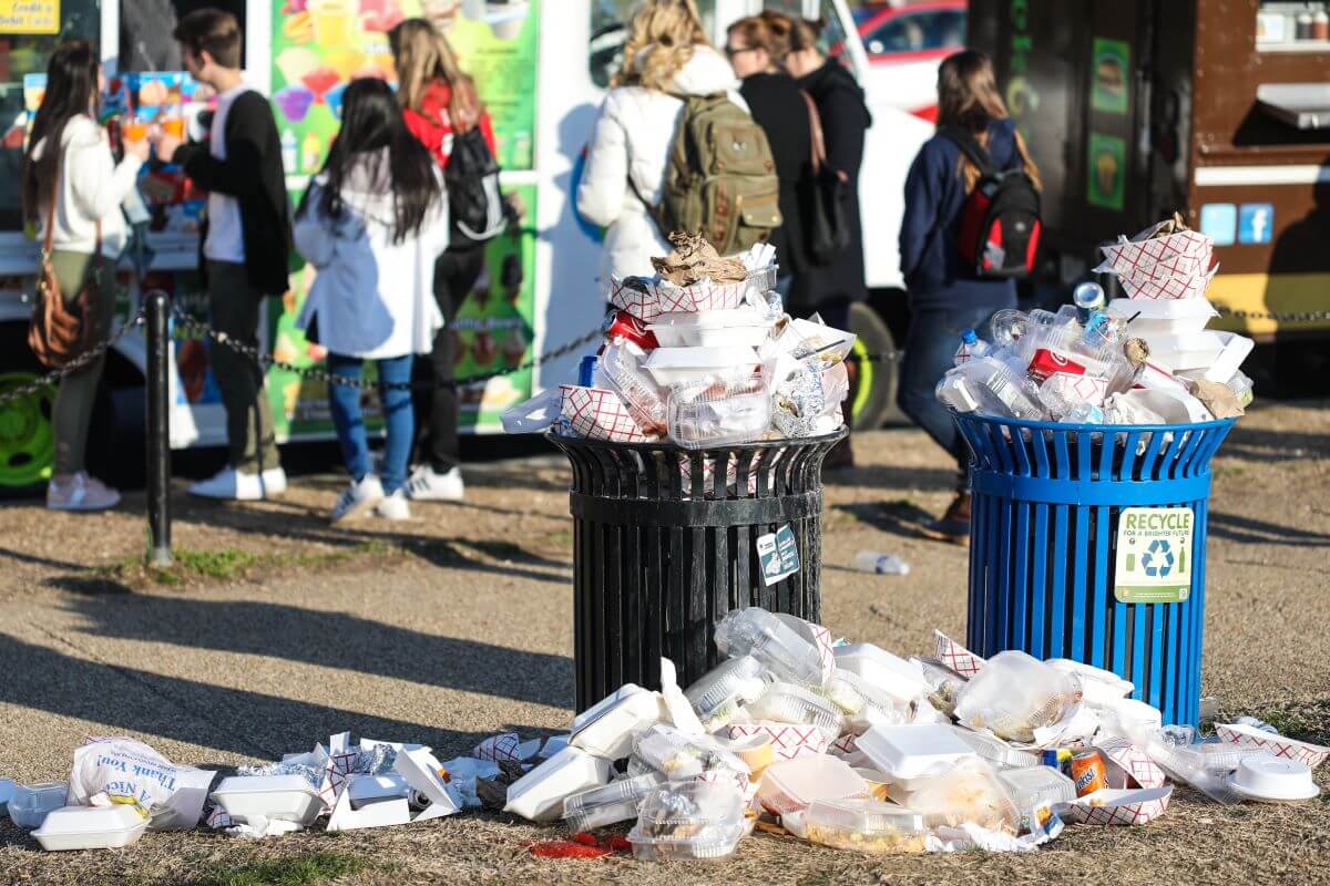 trash overflowing in two public receptacles with a line of people in the background