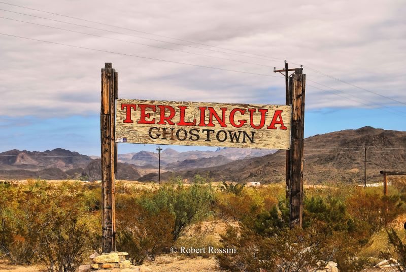 The entrance sign to Terlingua, describing it as a ghost town. The sign is surrounded by desert plants with mountains in the background.