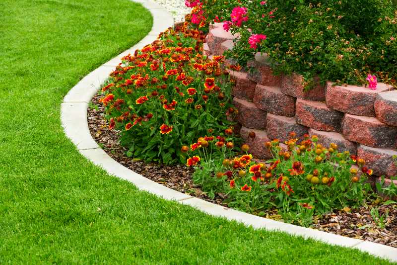 Curved concrete landscape edging separates lush green grass from a raised brick flower bed filled with vibrant red and yellow blooms.