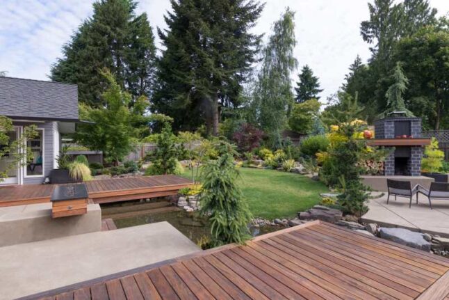 Back yard of a contemporary Pacific Northwest home featuring a deck a spanning creek-like water feature with a landscaped lawn and custom patio fireplace in the background.