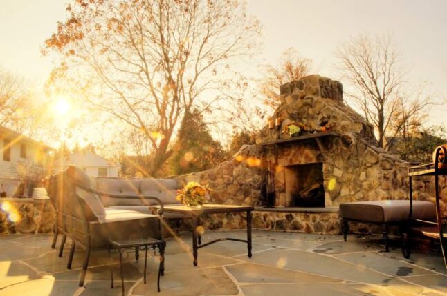 Low-angle view of a flagstone patio with an outdoor stone fireplace and furniture. Rays of sunlight stream down.