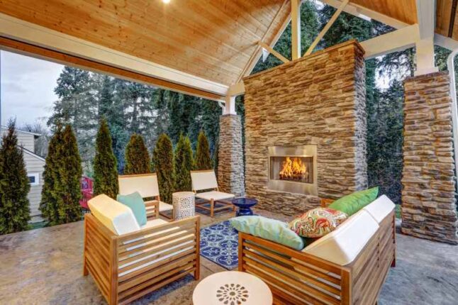 Chic covered back patio with built in gas fireplace, stone pillars, plank vaulted ceiling over cozy teak wood sofa set topped with white cushions and green pillows. Northwest, USA