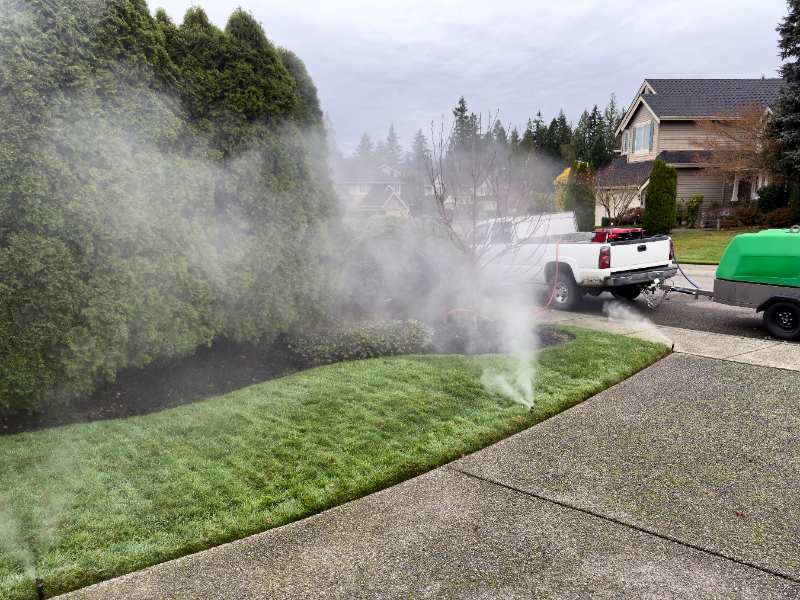 Worker winterizes residential lawn sprinkler system, mist spraying near shrubs, white service truck and green equipment trailer parked roadside today.
