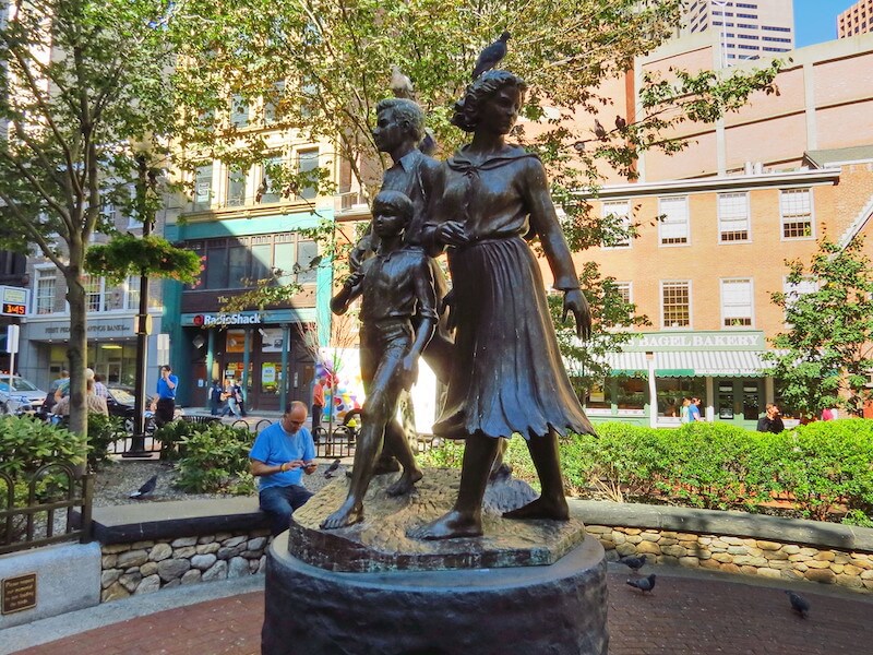 A statue of an Irish family of three, forming part of the Irish Famine Memorial along the Irish Heritage Trail, stands in the foreground surrounded by community members.