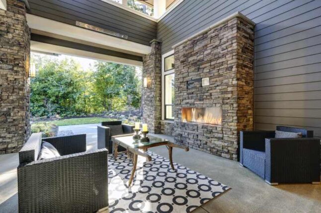 Well designed covered patio boasts stone fireplace, wicker patio chairs facing gorgeous rustic wood coffee table atop white and black geometric rug.