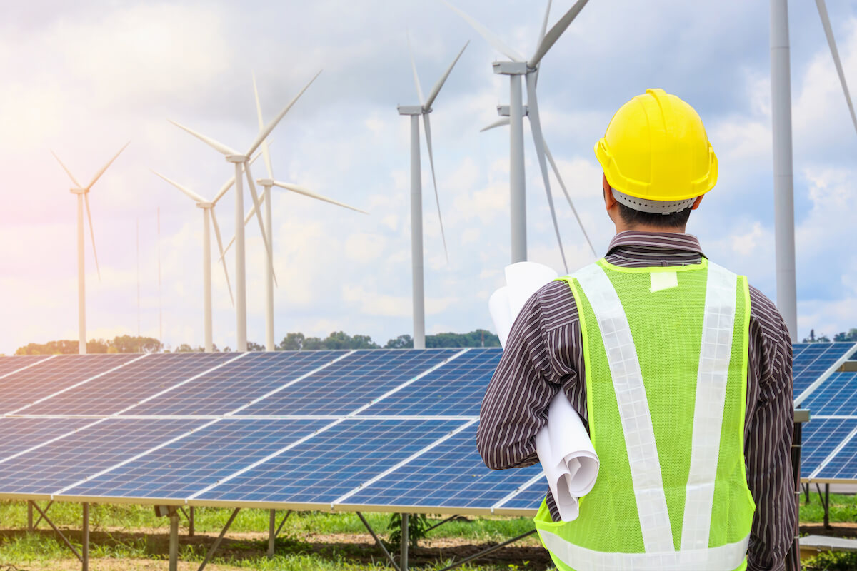 An engineer observes a row of solar panels and wind turbines in a grassy area.