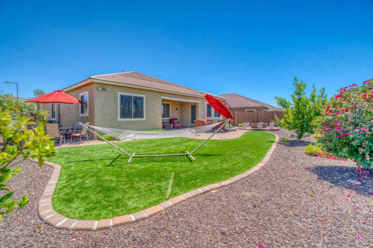 Spacious Arizona backyard with artificial grass, hammock, red umbrellas, patio furniture, desert landscaping, flowering shrubs, and single-story home exterior.