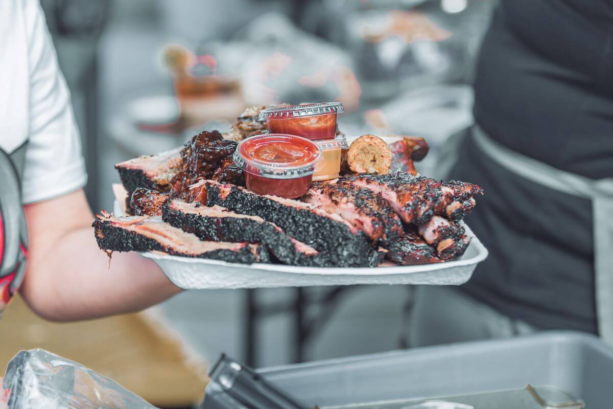 A person holds a tray piled with barbecue ribs and sausages, topped with containers of barbecue sauce.