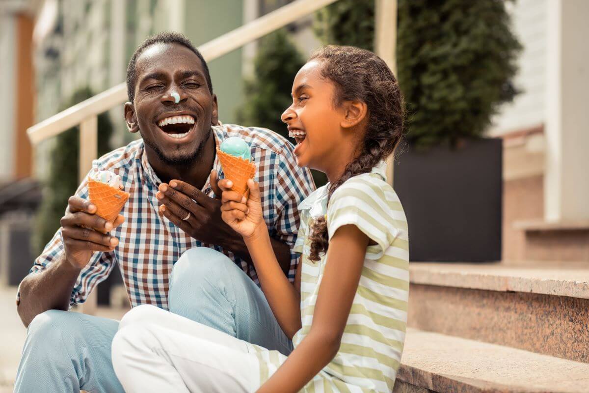 A father with ice cream on his nose laughs with his daughter while they sit together on a stoop eating ice cream cones.