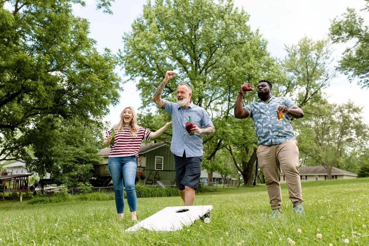Two men hold drinks and play cornhole in the backyard, while a female friend cheers them on.