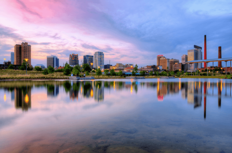 The colorful Birmingham skyline reflects in the water during a pink sunset.