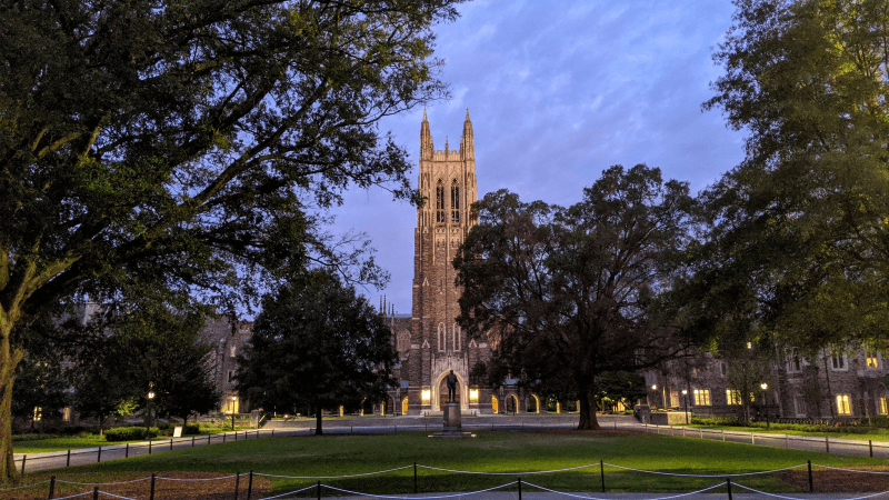 The iconic Duke University Chapel towers over a grassy yard with a statue and some trees as dusk falls on a cloudy evening in Durham.