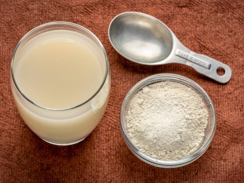 A glass of pale, cloudy diatomaceous earth mixed with water sits beside a tablespoon and a small glass bowl of white diatomaceous earth powder on a rust-colored textured surface.