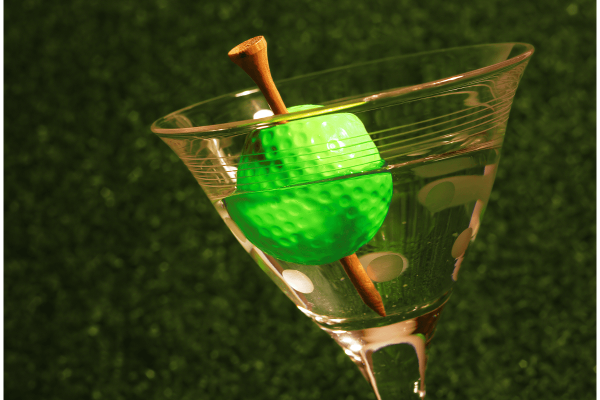 A martini glass, with a golf ball pierced by a tee to represent an olive on a toothpick, is set against a grass background.