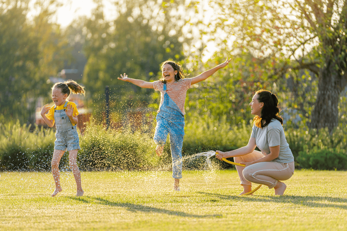 A mom playfully sprays her two young daughters with a hose in their large backyard