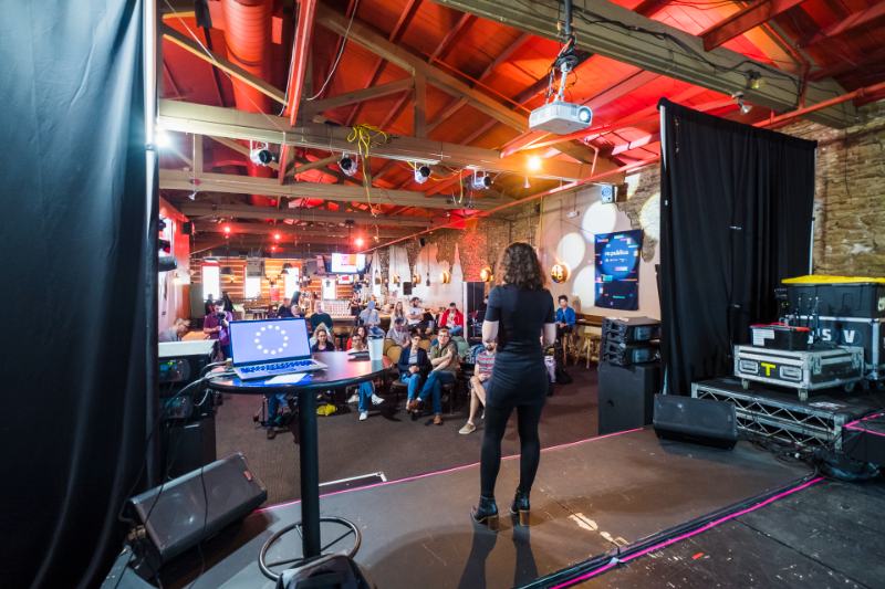 Speaker in a dark hoodie addresses a large SXSW crowd outdoors in Austin, with buses, buildings, and cloudy sky. 