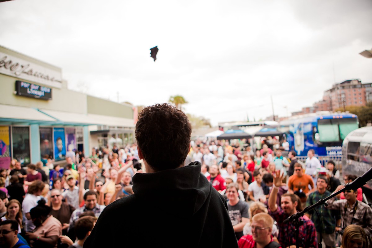 View from stage shows speaker or musician facing large outdoor crowd, hands raised, festival atmosphere with food trucks nearby today.