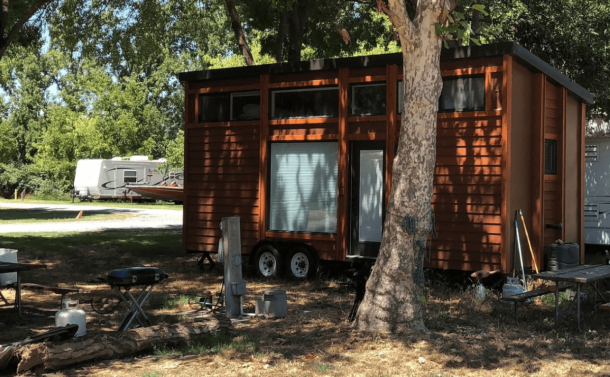 A shot of a brown tiny house on wheels parked on the grass behind a tree in the Sacramento River Delta in California