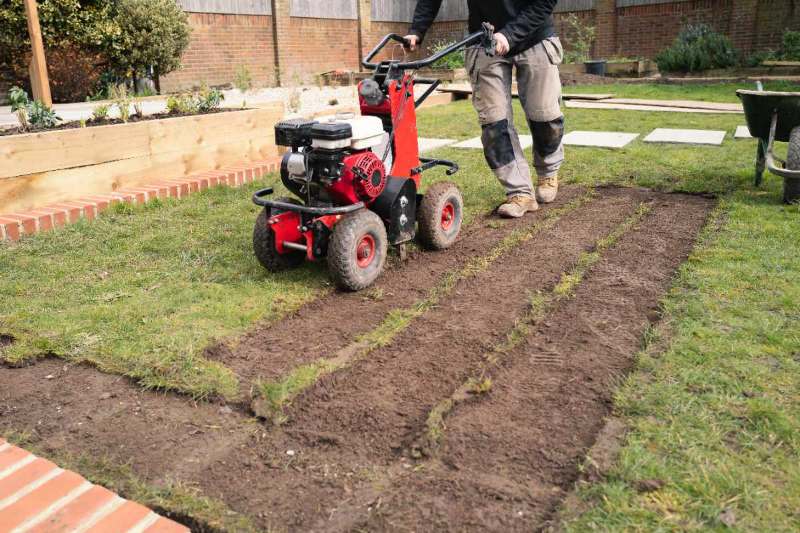 Worker using a red sod cutter to remove strips of grass from a lawn, exposing bare soil for landscaping
