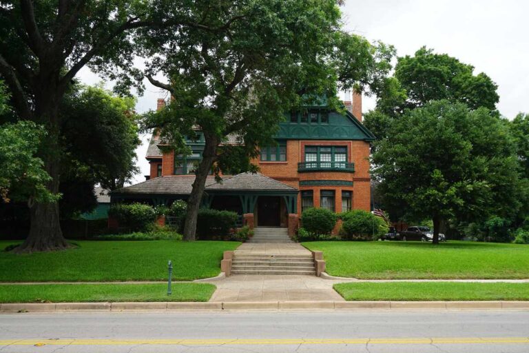 Lawn in front of a historic brick house in Waco, Texas, shaded by large trees and bordered by a quiet street.