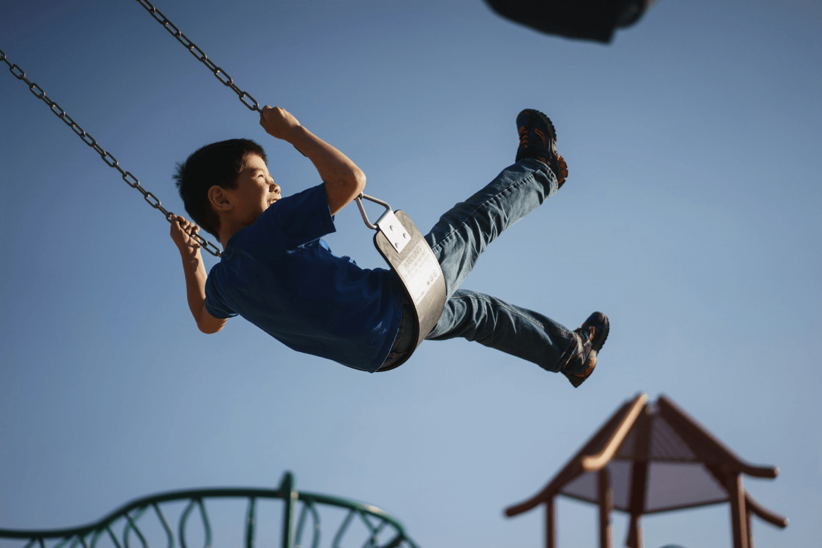 A young boy smiles while kicking his legs in the air on the swings at the playground