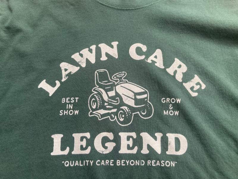 Green lawn care themed T-shirt featuring a vintage mower graphic, bold Legend text, and playful “Quality Care Beyond Reason” slogan.