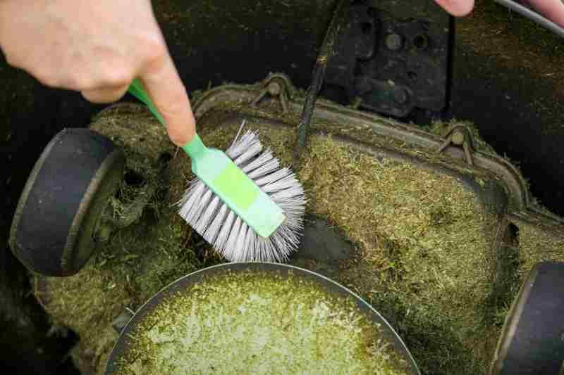 Close up view of man hand brushing off layer of wet grass stuck under a lawnmower