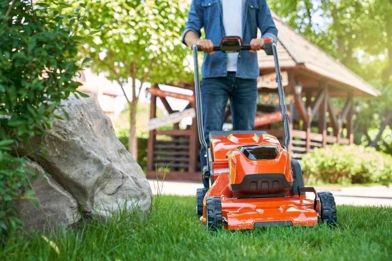 Male gardener in denim clothes using lawn mower on backyard
