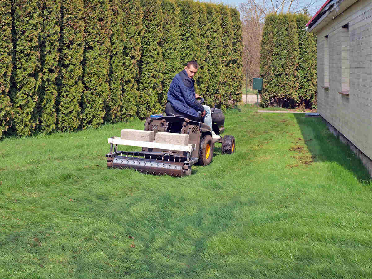 A man on a ride-on mower is using a pull-behind core aerator on a lawn.