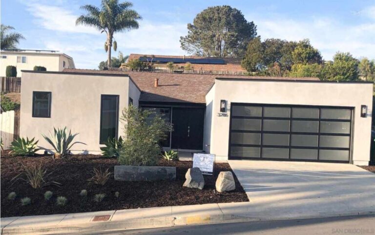 Modern single-story San Diego home with flat roof, black-framed garage door, drought-tolerant landscaping, wide driveway, and palm trees behind.