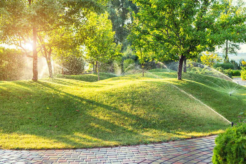 Automatic garden watering system with different sprinklers installed under turf. Landscape design with lawn hills and fruit garden irrigated with smart autonomous sprayers