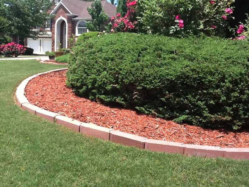 Neatly mulched garden bed with red wood chips bordered by curved stone edging in a well-maintained front yard with green lawn, shrubs, and blooming flowers.